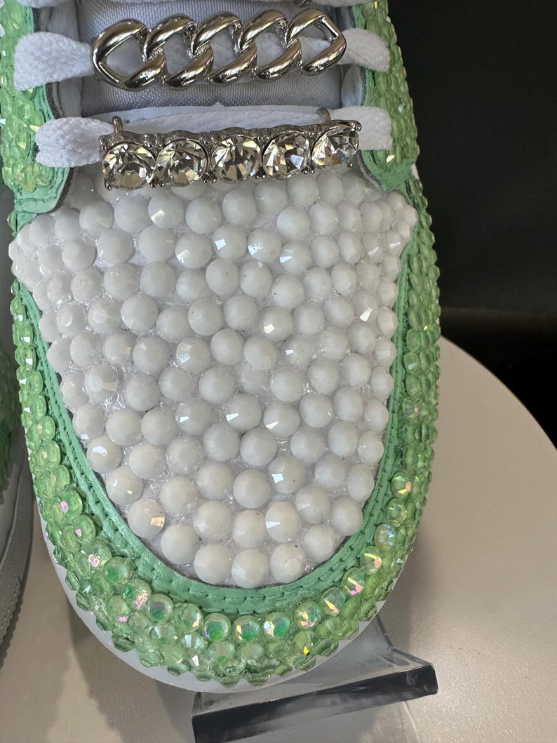 May include: Close-up of a white sneaker with a green trim. The sneaker is covered in white and green rhinestones. A silver chain and a row of clear rhinestones are on the laces.