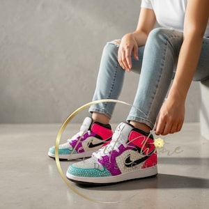 May include: A pair of bedazzled high-top sneakers with a white base, featuring teal, purple, and pink accents. The sneakers have a black Nike swoosh and are embellished with sparkling rhinestones. The person is wearing light blue jeans.