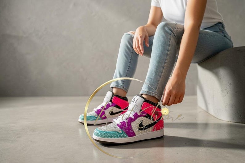 May include: A pair of bedazzled high-top sneakers with a white base, featuring teal, purple, and pink accents. The sneakers have a black Nike swoosh and are embellished with sparkling rhinestones. The person is wearing light blue jeans.
