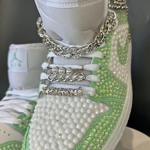 May include: White high-top sneakers with green accents and rhinestones. The sneakers have a silver chain and pearl accents on the laces. The text "AIR" is visible on the shoe.
