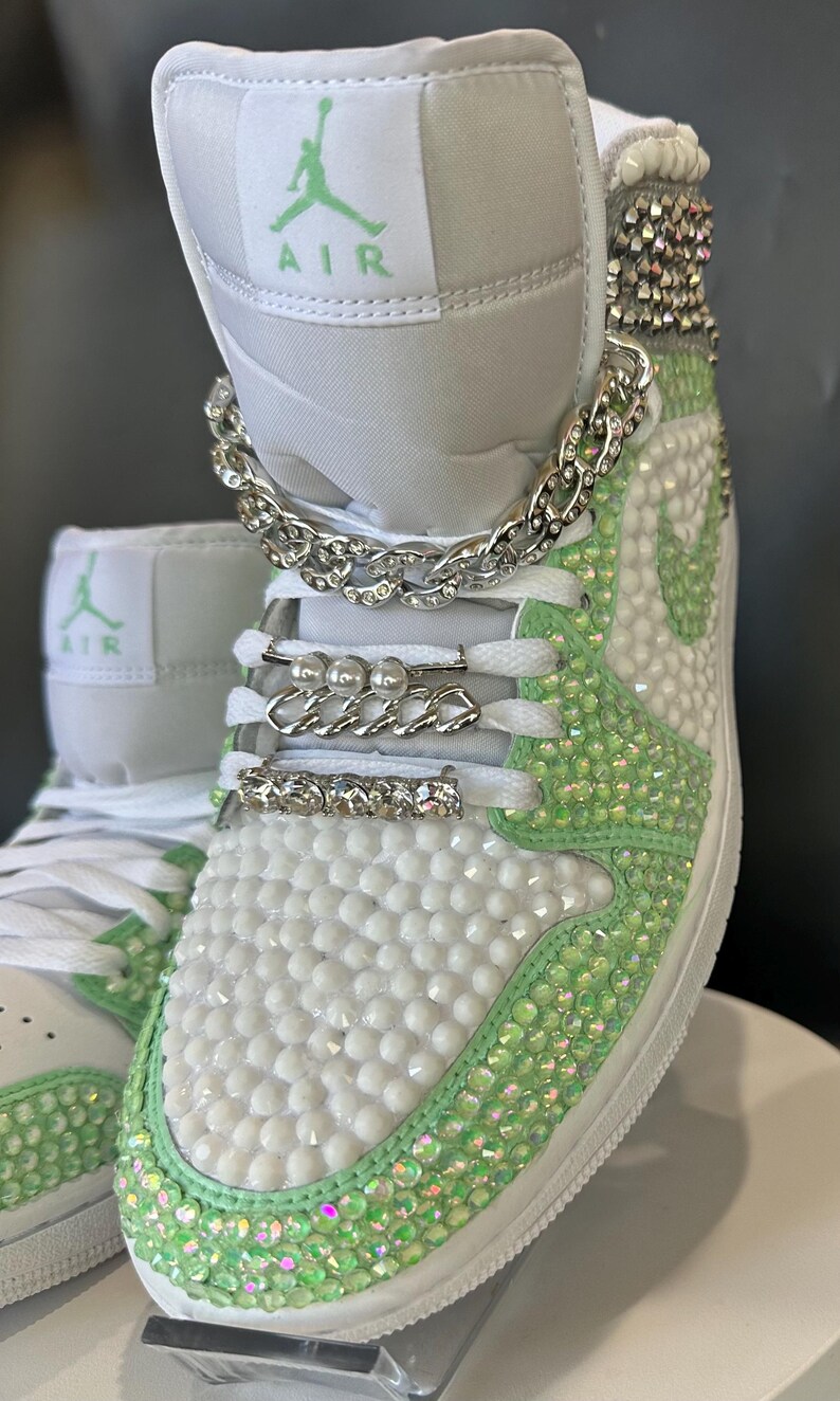 May include: White high-top sneakers with green accents and rhinestones. The sneakers have a silver chain and pearl accents on the laces. The text "AIR" is visible on the shoe.
