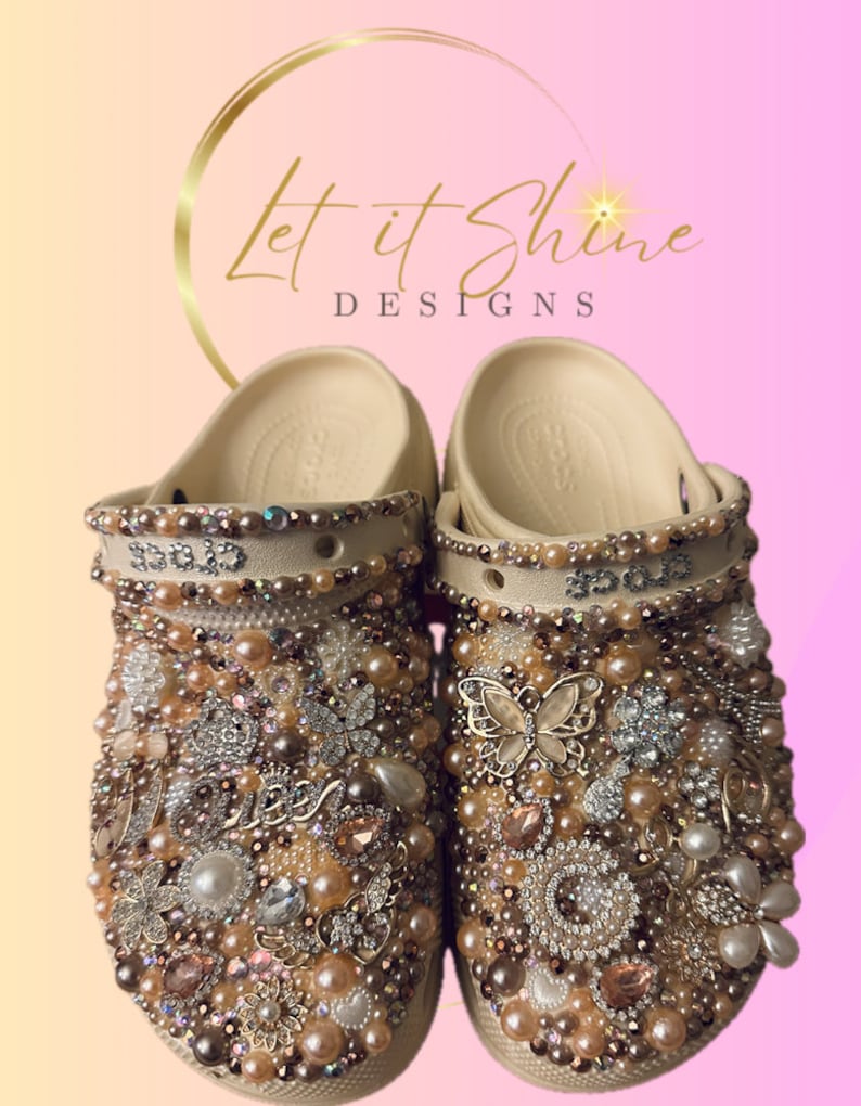 Pu&ograve; includere: Un paio di zoccoli beige decorati con perle, strass e dettagli dorati. Gli zoccoli sono adornati con un ciondolo a forma di farfalla e la parola "Crocs" &egrave; visibile sul lato.