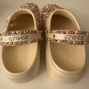Pu&ograve; includere: Un paio di zoccoli Crocs bianchi decorati con perline rosa e iridescenti. Gli zoccoli hanno un cinturino sulla parte superiore con la parola "crocs" scritta in strass. Le suole sono contrassegnate "iconic comfort crocs".