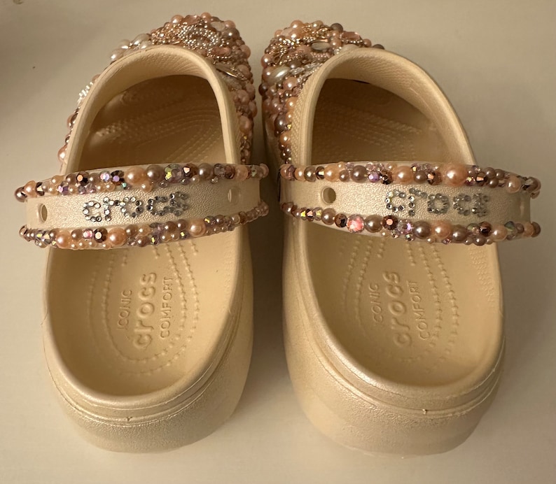 Pu&ograve; includere: Un paio di zoccoli Crocs bianchi decorati con perline rosa e iridescenti. Gli zoccoli hanno un cinturino sulla parte superiore con la parola "crocs" scritta in strass. Le suole sono contrassegnate "iconic comfort crocs".