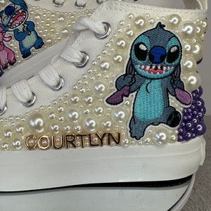 May include: White canvas high-top sneakers with a blue Stitch character embroidered on the side. The shoes are decorated with white pearls and have a gold name "COURTLYN" on the side.