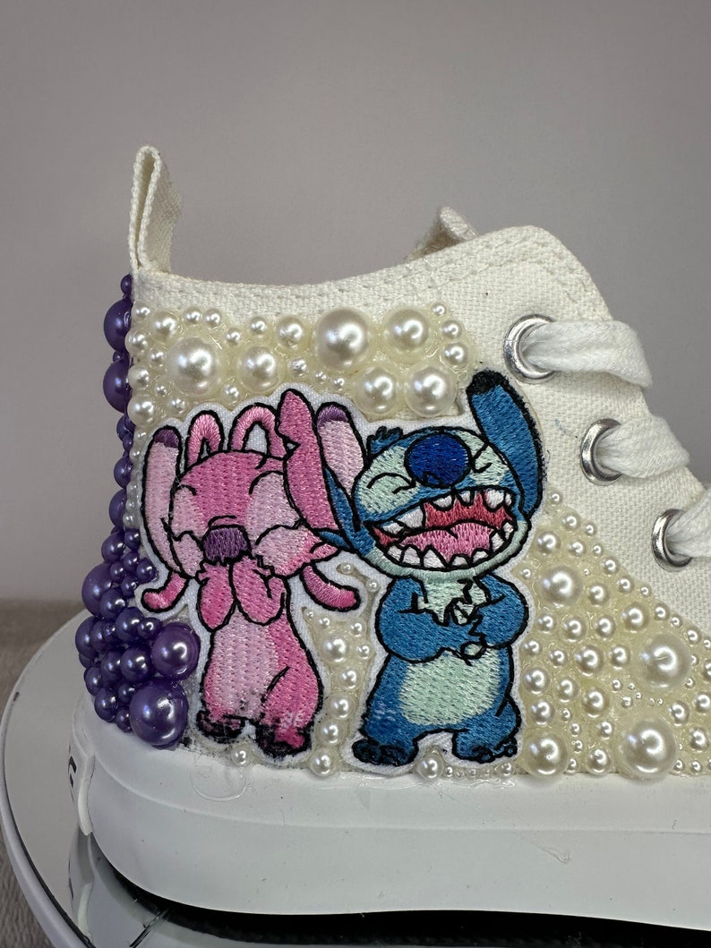 May include: White high-top sneakers with a pearl embellishment and embroidered images of Stitch and Angel from the Disney movie Lilo & Stitch.