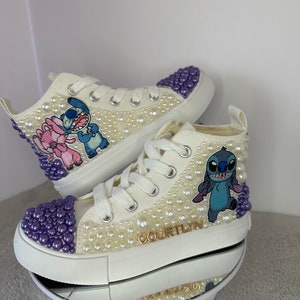 May include: White high-top sneakers with purple and white pearl embellishments. The sneakers feature an embroidered Stitch character and the name "COURTLYN" in gold lettering.