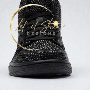 May include: Black high-top sneaker covered in small, sparkling embellishments. The shoe features black laces and a black sole. The text "Let it Shine DESIGNS" is visible in a gold circular design.
