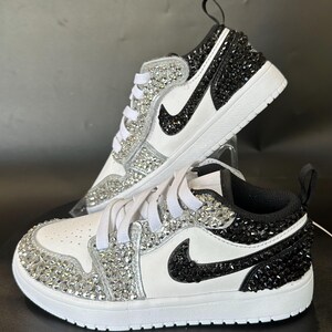 Puede incluir: Zapatillas blancas de ca&ntilde;a baja con pedrer&iacute;a negra y plateada. El logotipo de swoosh es negro con pedrer&iacute;a plateada.