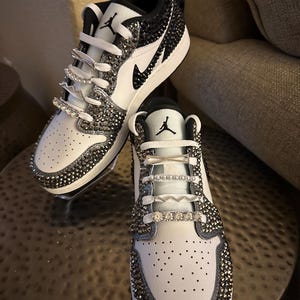 Puede incluir: Zapatillas bajas blancas y negras con un logotipo Swoosh negro y pedrer&iacute;a que cubre toda la zapatilla.