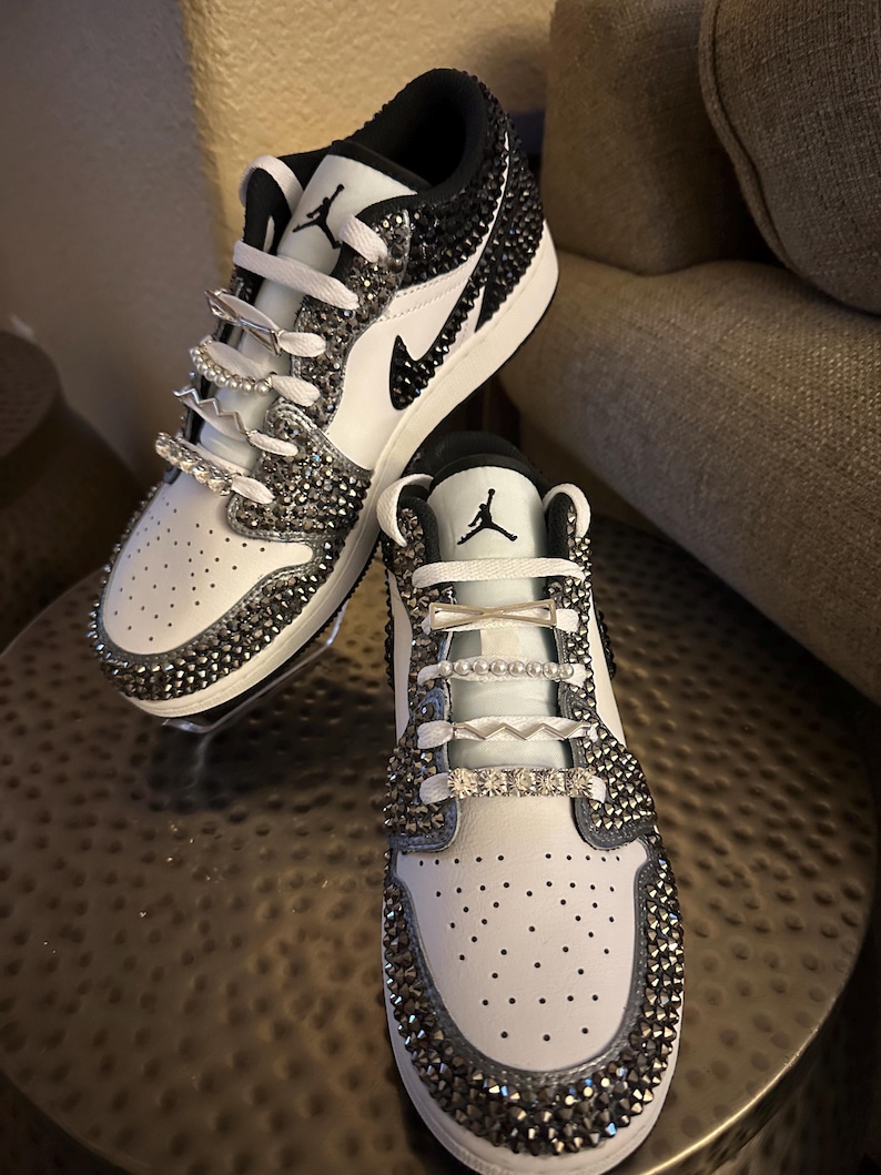 Puede incluir: Zapatillas bajas blancas y negras con un logotipo Swoosh negro y pedrer&iacute;a que cubre toda la zapatilla.