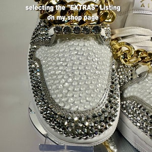 May include: White sneakers with a gold chain and rhinestones. The sneakers have a white leather upper with a gold chain around the ankle. The sneakers are covered in rhinestones. The text "Add lace charms to your order by selecting the "EXTRAS" Listing on my shop page" is written on the image.