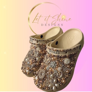 Pu&ograve; includere: Zoccoli beige decorati con perle, strass e altri abbellimenti. Gli zoccoli sono ricoperti da diverse tonalit&agrave; dorate, argentate e rosa. Il testo "Let it Shine Designs" &egrave; visibile sullo sfondo.