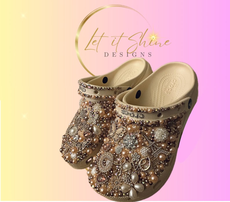 Pu&ograve; includere: Zoccoli beige decorati con perle, strass e altri abbellimenti. Gli zoccoli sono ricoperti da diverse tonalit&agrave; dorate, argentate e rosa. Il testo "Let it Shine Designs" &egrave; visibile sullo sfondo.