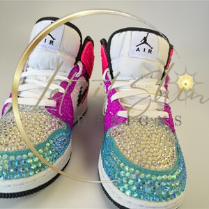 May include: A pair of bedazzled high-top sneakers. The toe and sides are covered in iridescent rhinestones, transitioning from teal to silver. The upper features white, pink, and black accents, with white laces and the word "AIR" visible on the tongue.