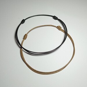 May include: Two minimalist bracelets. One is black, the other is tan. Both are made of thin, woven cord and feature adjustable sliding knots. The bracelets are simple and elegant, suitable for everyday wear.