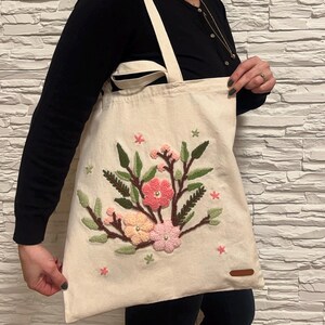 Op de afbeelding: Een witte canvas tote bag met een geborduurd bloemenmotief. De bloemen zijn roze, perzikkleurig en wit met groene bladeren. De tas heeft bruine hengsels.