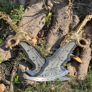 May include: Two decorative metal axes with gold-colored dragon-shaped handles and silver blades. The blades feature etched designs and runes. The axes are displayed on a tree trunk with green foliage.
