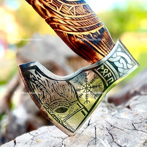 May include: A silver axe head with a detailed design of a wolf and a compass with runes. The axe head is attached to a wooden handle with a carved design.