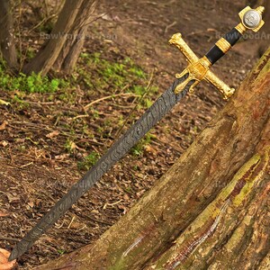 May include: A gold-plated sword with a black handle and a damascus steel blade. The sword is resting on a tree trunk in a woodland setting.