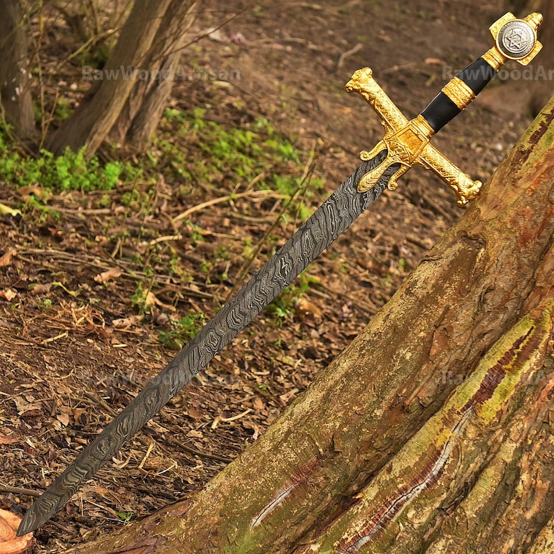 May include: A gold-plated sword with a black handle and a damascus steel blade. The sword is resting on a tree trunk in a woodland setting.