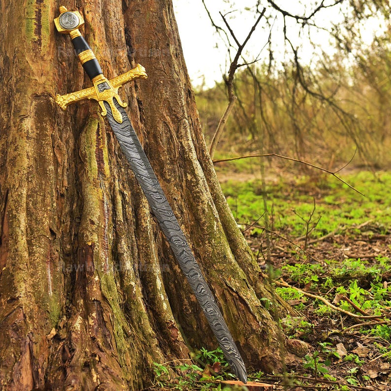 May include: A sword with a gold handle and a black blade, featuring intricate details, is resting against a tree trunk in a woodland setting.