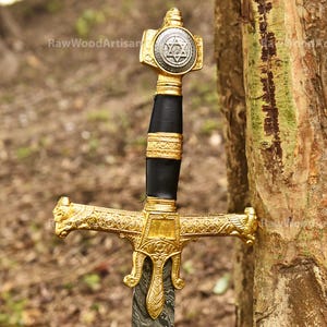 May include: A gold-coloured sword with a black handle and a Star of David on the pommel. The sword is resting against a tree trunk.