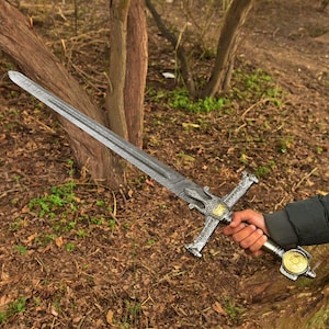 Op de afbeelding: Een gedetailleerd zwaard met een patroon op het lemmet en een sierlijke pareerstang. De handgreep heeft een goudkleurig medaillon. Het zwaard wordt vastgehouden tegen een achtergrond van bomen en bladeren, wat een buitenomgeving suggereert.