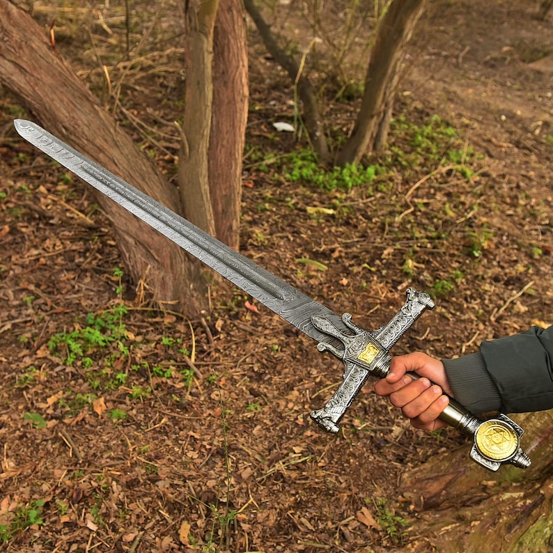 Op de afbeelding: Een gedetailleerd zwaard met een patroon op het lemmet en een sierlijke pareerstang. De handgreep heeft een goudkleurig medaillon. Het zwaard wordt vastgehouden tegen een achtergrond van bomen en bladeren, wat een buitenomgeving suggereert.