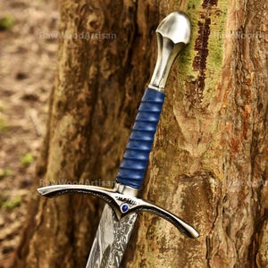May include: A sword with a patterned Damascus steel blade, a blue leather-wrapped grip, and a polished silver-toned pommel. The crossguard features a small blue gemstone at the center. The text RawWoodArtisan is visible on the blade.