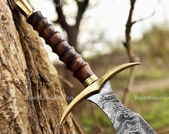 May include: A sword with a wooden handle and a gold-colored hilt. The blade is made of Damascus steel and has a wavy pattern. The sword is resting against a tree trunk.