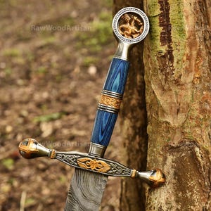May include: A decorative sword with a blue and gold handle and a patterned blade. The handle features a gold dragon emblem and intricate gold detailing. The sword is leaning against a tree trunk.