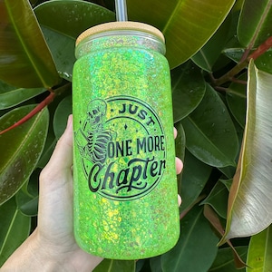 May include: A green glitter tumbler with a bamboo lid and straw. The tumbler features the text "Just One More Chapter" with a skeleton reading a book. The background is lush green foliage.