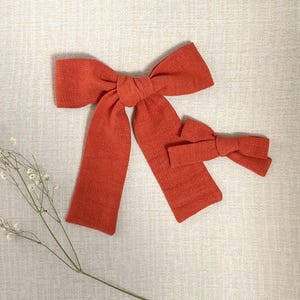 May include: Two burnt orange fabric bows. A large bow with long tails and a smaller matching bow. The bows are made of a textured fabric, set against a neutral background.