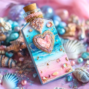May include: A decorative glass bottle with a cork stopper, adorned with a heart-shaped embellishment. The bottle features a beach-themed design with pink and blue hues, embellished with beads and glitter. The bottle is surrounded by seashells and pearls.