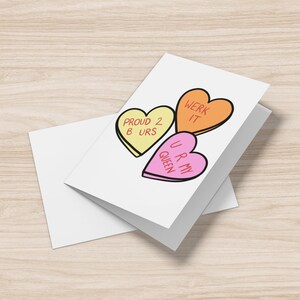 May include: A white greeting card featuring three candy heart illustrations. The hearts are yellow, orange, and pink, each with a different message: "PROUD 2 B URS", "WERK IT", and "U R MY QUEEN". A white envelope is next to the card.