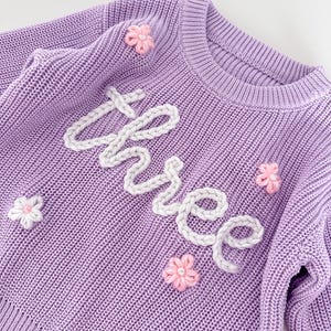 Peut inclure: Un pull en tricot lilas avec le mot "three" brodé en fil blanc. Le pull est orné de petites fleurs roses et blanches. Ce vêtement pour enfant présente un col rond et est de couleur unie.