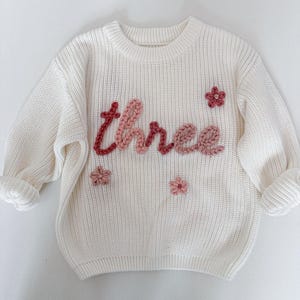 Peut inclure: Pull en tricot crème avec le mot "three" en fil rose et rouge texturé. Le pull présente de petits motifs de fleurs brodées dans des tons de rose. Un vêtement confortable et élégant pour l'anniversaire d'un enfant.