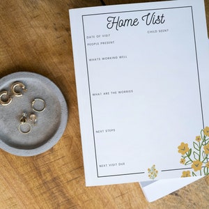 May include: A white sheet of paper with a black border and the text "Home Vist" in a handwritten font. The paper has sections for "Date of Visit", "People Present", "What's Working Well", "What are the Worries", "Next Steps", and "Next Visit Due". There is a small floral design in the bottom right corner.