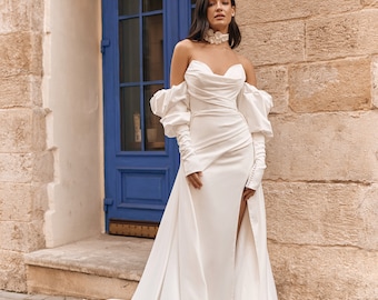 May include: A white off-the-shoulder wedding dress with a fitted bodice, a flowing skirt, and long, puffy sleeves. The dress has a thigh-high slit and a delicate white flower choker.