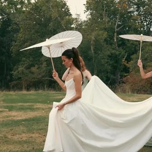 K&ouml;nnte beinhalten: Eine Braut in einem wei&szlig;en Hochzeitskleid mit langer Schleppe geht &uuml;ber eine Wiese und h&auml;lt das Kleid. Sie wird von anderen begleitet, die wei&szlig;e Sonnenschirme halten. Der Hintergrund zeigt B&auml;ume und einen bew&ouml;lkten Himmel.