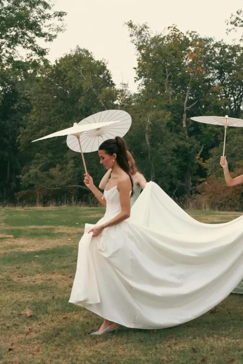 K&ouml;nnte beinhalten: Eine Braut in einem wei&szlig;en Hochzeitskleid mit langer Schleppe geht &uuml;ber eine Wiese und h&auml;lt das Kleid. Sie wird von anderen begleitet, die wei&szlig;e Sonnenschirme halten. Der Hintergrund zeigt B&auml;ume und einen bew&ouml;lkten Himmel.