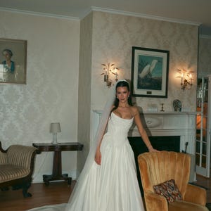 K&ouml;nnte beinhalten: Ein tr&auml;gerloses wei&szlig;es Brautkleid mit eng anliegendem Oberteil und flie&szlig;endem Rock. Die Braut tr&auml;gt einen Schleier. Der Raum hat antike M&ouml;bel und einen Kamin. Der Raum hat eine Vintage-&Auml;sthetik.