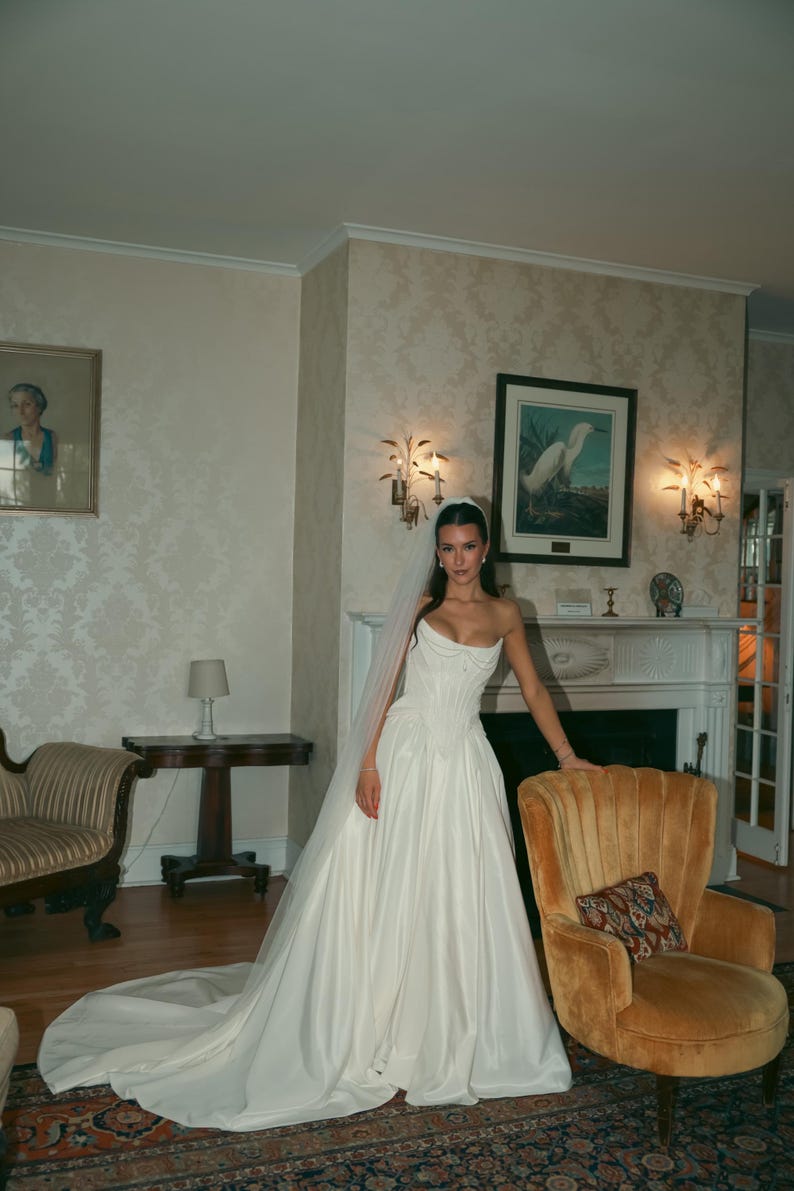 K&ouml;nnte beinhalten: Ein tr&auml;gerloses wei&szlig;es Brautkleid mit eng anliegendem Oberteil und flie&szlig;endem Rock. Die Braut tr&auml;gt einen Schleier. Der Raum hat antike M&ouml;bel und einen Kamin. Der Raum hat eine Vintage-&Auml;sthetik.