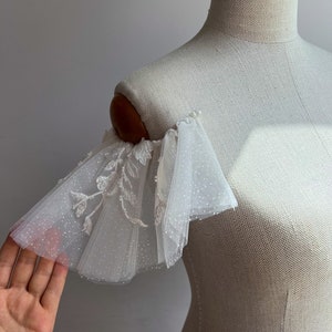 May include: White tulle bridal shoulder sleeves with delicate floral embroidery and a subtle sparkle. The sleeves are gathered and ruffled, creating a romantic and ethereal look.