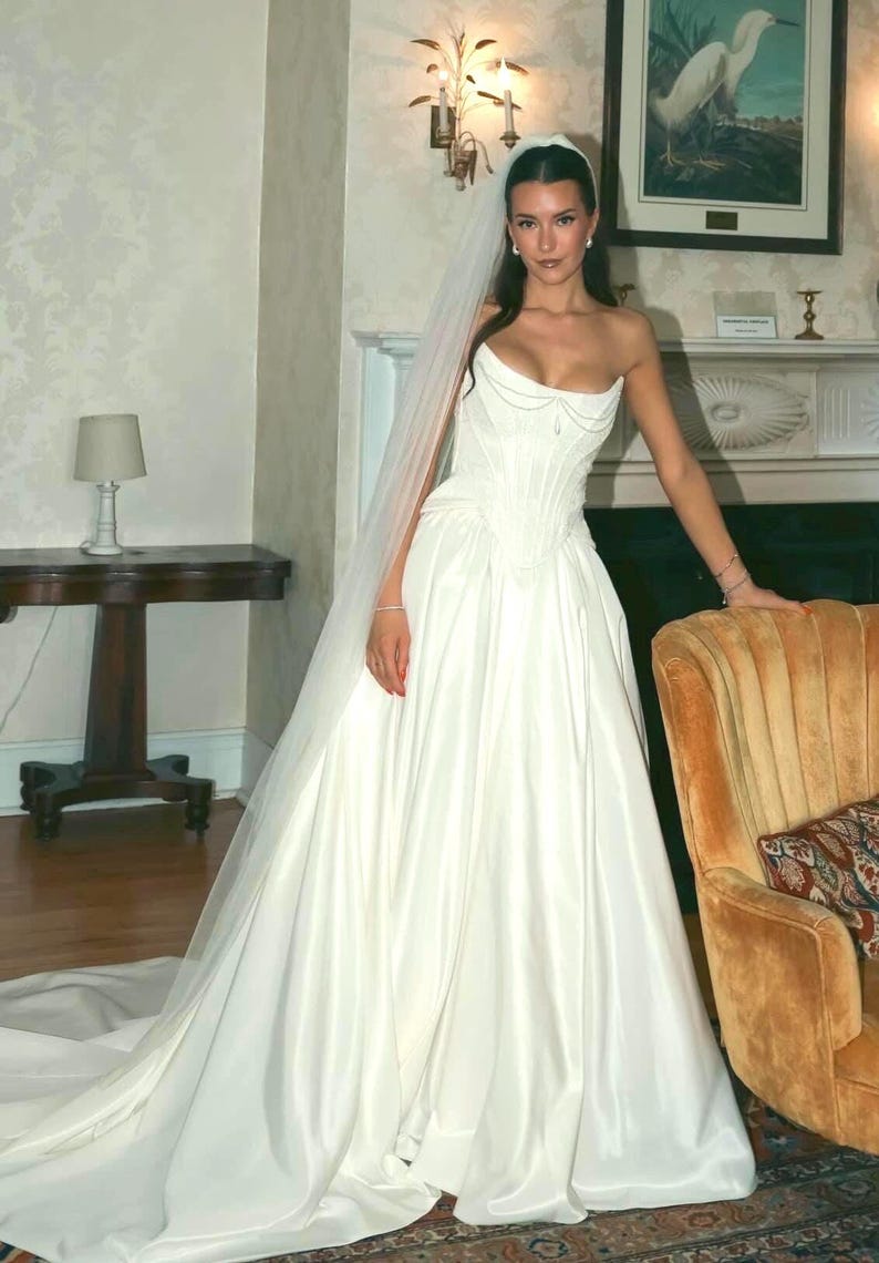 K&ouml;nnte beinhalten: Ein wei&szlig;es, tr&auml;gerloses Hochzeitskleid mit einem eng anliegenden Oberteil und einem weiten Rock. Das Kleid hat einen herzf&ouml;rmigen Ausschnitt und eine lange Schleppe. Ein durchsichtiger Schleier ist &uuml;ber dem Kopf drapiert.