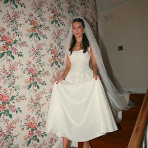 K&ouml;nnte beinhalten: Eine Braut in einem wei&szlig;en Satin-Hochzeitskleid mit herzf&ouml;rmigem Ausschnitt und flie&szlig;endem Rock geht eine Treppe hinunter. Sie tr&auml;gt einen Schleier und hellblaue Abs&auml;tze. Der Hintergrund zeigt eine florale Tapete und eine rot ausgelegte Treppe.