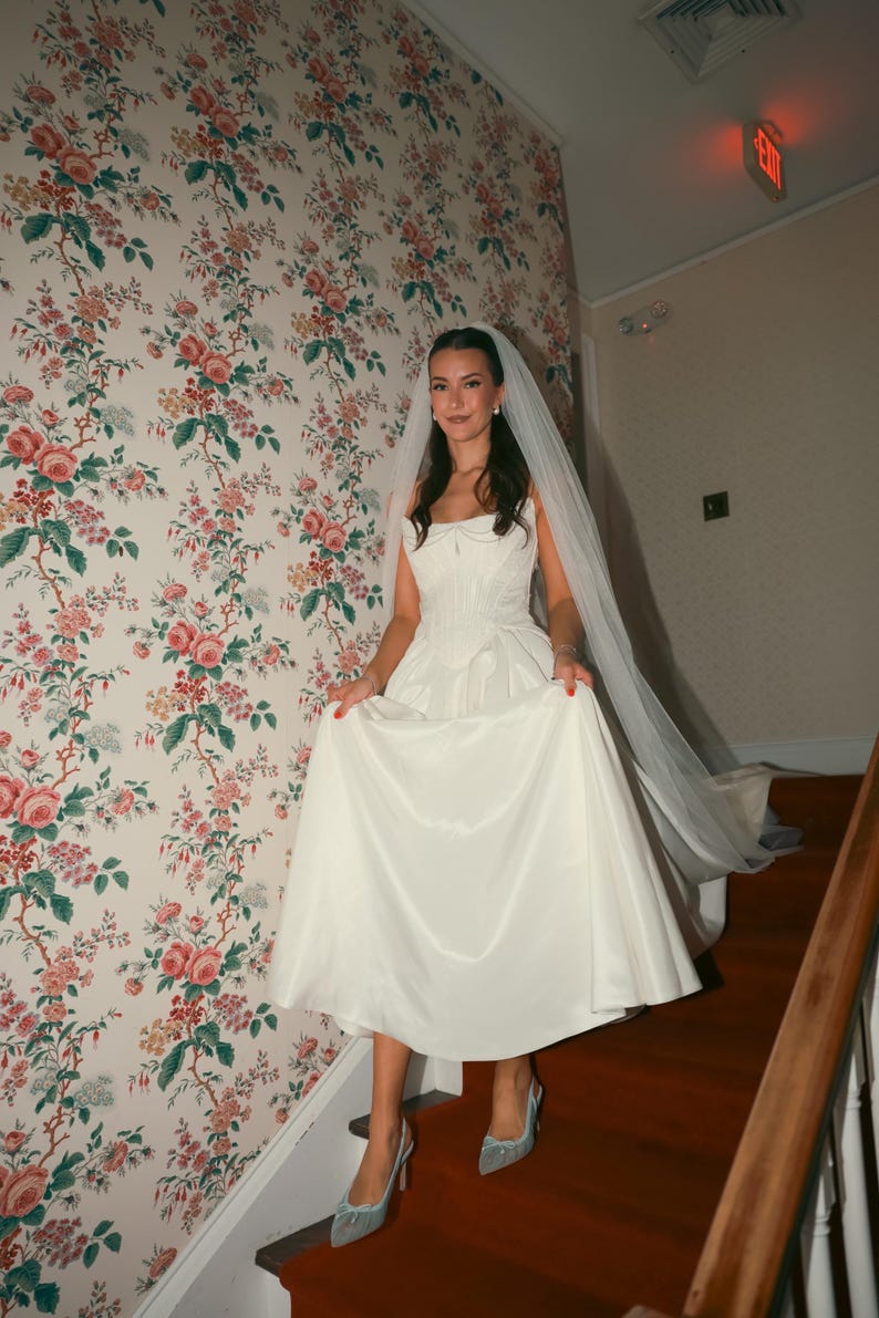 K&ouml;nnte beinhalten: Eine Braut in einem wei&szlig;en Satin-Hochzeitskleid mit herzf&ouml;rmigem Ausschnitt und flie&szlig;endem Rock geht eine Treppe hinunter. Sie tr&auml;gt einen Schleier und hellblaue Abs&auml;tze. Der Hintergrund zeigt eine florale Tapete und eine rot ausgelegte Treppe.