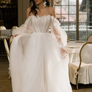 May include: A white off-the-shoulder wedding dress with a fitted bodice and a flowing tulle skirt. The dress has delicate floral embellishments and sheer puff sleeves.