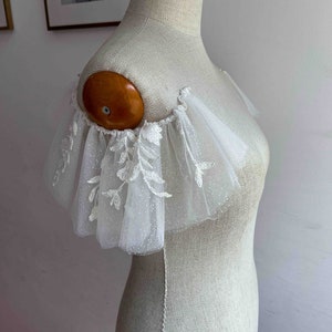 May include: White tulle bridal shoulder piece with delicate floral embroidery and a scalloped edge.